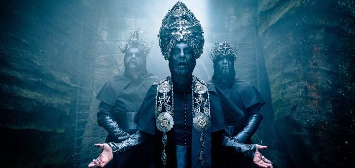 Bandfoto von Behemoth. Der Sänger steht vorne mittig mit Segnungsgestus in einer Art verkehrten Bischofskleidung. Zwei weitere Musiker stehen hinter ihm in einer andächtigen Pose und in ähnlich gestaltetem "Ornat".