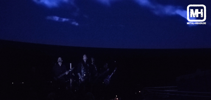 Hallig live im Planetarium Bochum. Die untere Hälfte des Bildes ist schwarz. Darin sind die Silhouetten der Band erkennbar. Die obere Bildhälfte zeigt die Lichtshow mit einem dunkelblauen Gewitterhimmel.