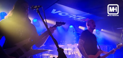 White Ward live in Siegen. Aufnahme der Band auf der Bühne. Vorne links einer der Gitarristen, rechts der Sänger und Bassist. Im Hintergrund sieht man das Schlagzeug. Alles in kaltem, blauen Licht. Ein Scheinwerfer blendet in die Kamera.