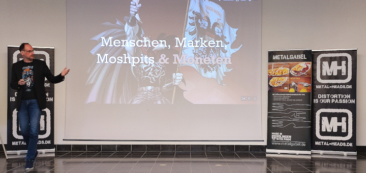 Menschen, Marken, Moshpits: René Thaller steht neben seiner Präsentation und zeigt auf diese. Rechts davon stehen zwei Roll-Up-Displays, eines von Metalgabel und eines von Metal-Heads.de. Ein Roll-Up-Display wird vom Referenten verdeckt.