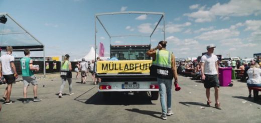 Blick von hinten auf einen Pritschenwagen mit der Aufschrift Müllabfuhr. Das Fahrzeug fährt auf einem Festivalgelände neben Biertischgarnituren vorbei. Mitglieder der Festivalcrew begleiten das Fahrzeug zu Fuß.