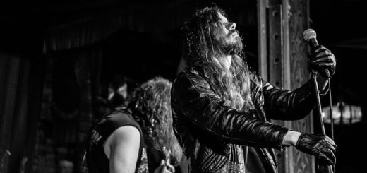 Black Sorcery live. Der Sänger mit schwarzen Handschuhen und Lederjacke steht mit geschlossenen Augen am Mikro. Im Hintergrund ist ein weiterer Musiker von hinten zu sehen.