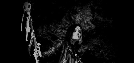 Witch N. von Ashtar aus ein wenig Abstand fotografiert, mit dunkel geschminkten Augen und einem Symbol auf der Stirn. Sie Musikerin blickt in die Ferne. In der Hand hält sie einen Stab mit verschiedenen Tierknochen und -schädeln.