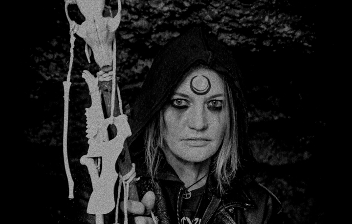 Witch N. von Ashtar mit dunkel geschminkten Augen und einem Symbol auf der Stirn blickt in die Kamera. In der Hand hält sie einen Stab mit verschiedenen Tierknochen und -schädeln.