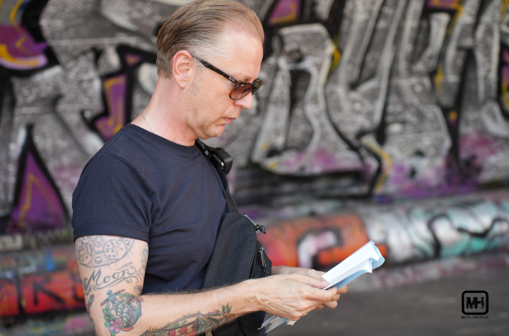 Vainstream 2023: Festival Director Tom Naber, mit Sonnenbrille und Funkgerät, prüft kritisch ein paar Unterlagen. Im Hintergrund eine mit Graffiti besprühte Wand.