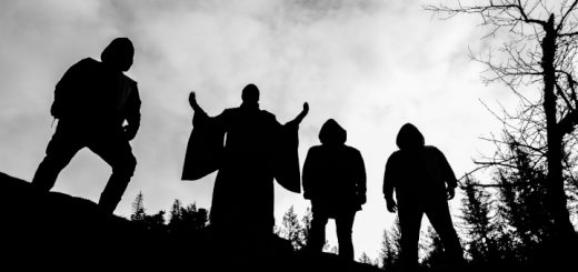 Die Band Uada steht auf einer Anhöhe vor ein paar abgestorbenen Bäumen. Das Bild ist mit starkem Gegenlicht aufgenommen, so dass die Bandmitglieder nur in Umrissen erkennbar sind.