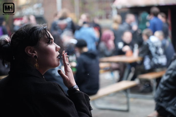 Culthe Fest 2024: Impressionen. Im Vordergrund rauch eine schwarz gekleidete Frau eine Zigarette. Im Hintergrund ist unscharf das Publikum erkennbar.