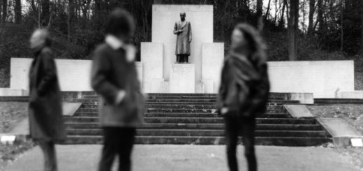 Die Bandmitglieder von FLUISTERAARS vor einem Denkmal. Die Bandmitglieder sind unscharf gestellt, während im Hintergrund die Statue des Denkmals scharf gestellt ist. Zwei Bandmitglieder blicken in die Ferne, eines zur Statue.