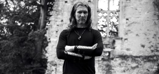 The Watcher vor einer alten Ruine. Der Solomusiker von Fellwarden hat die Arme verschränkt und blickt ernst in die Kamera.