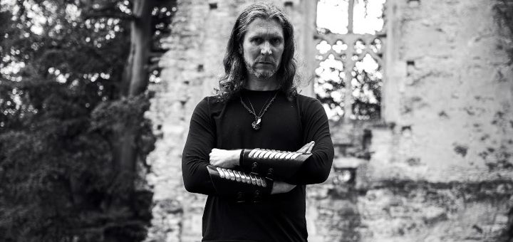 The Watcher vor einer alten Ruine. Der Solomusiker von Fellwarden hat die Arme verschränkt und blickt ernst in die Kamera.