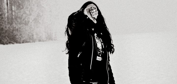 Der Solomusiker von Uprising steht in einer Schneelandschaft vor einer Baumreihe. Das Gesicht verdeckt seine Hand, die eine Geste mit dem Daumen nach unten bildet.