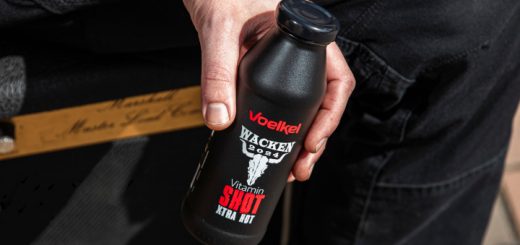 Jemand sitzt auf einem Verstärker und hält einen schwarzen Voelkel Wacken Ingwer Shot in der Hand.