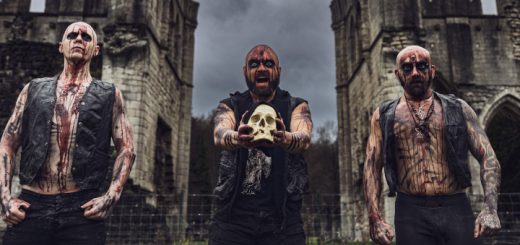 TRIVAX stehen mit Kunstblut überströmt in einer alten Ruine. Der Sänger hält einen Schädel in die Kamera und zieht Grimassen.