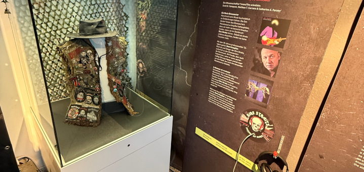 Eine Metal-Kutte in einem Schaukasten, daneben Informationen an der Wand mit einem Kopfhörer.