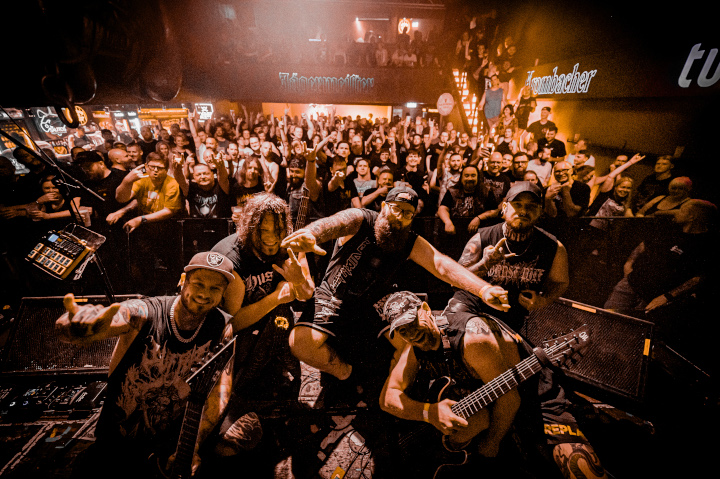 Die Band beim Gruppenfoto auf der Bühne mit dem Rücken zum Publikum, das im Hintergrund jubelt.