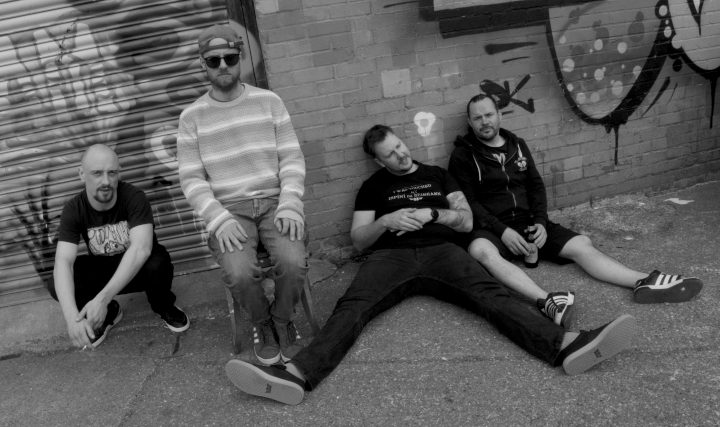 Bandmitglieder von Elephant Tree lungern vor einer Wand und einem Tor mit Grafitti herum.